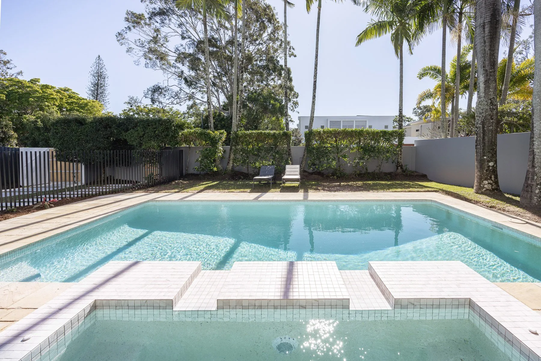 An outdoor swimming pool is surrounded by tiled decking, lounge chairs, and tropical greenery.