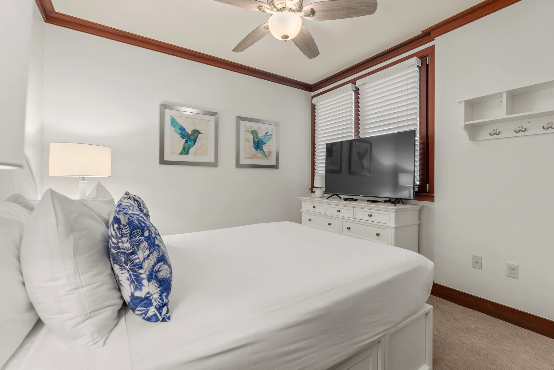 Un dormitorio luminoso con una cama blanca, cuadros enmarcados y un televisor junto a la ventana.