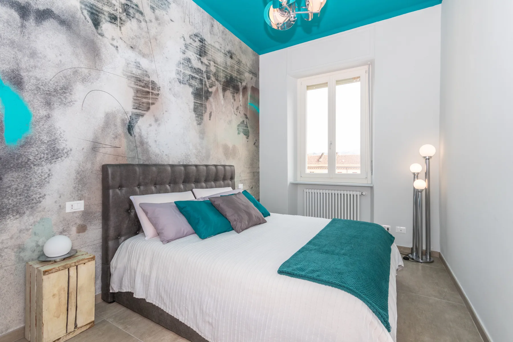 A bedroom with a bed dressed in white and teal bedding beneath a teal ceiling.