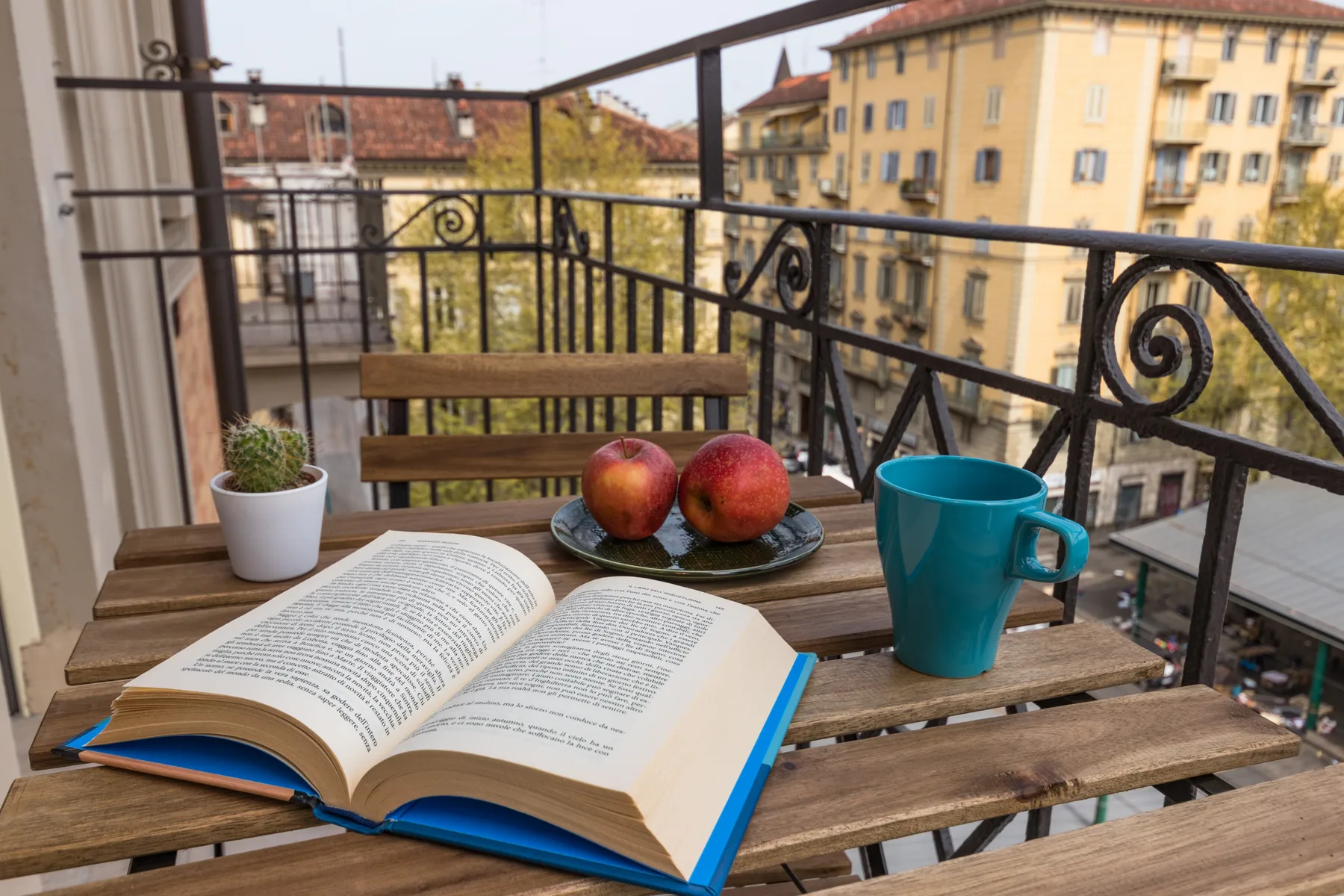 A balcony table holds an open book, fruit, a plant, and a blue mug.