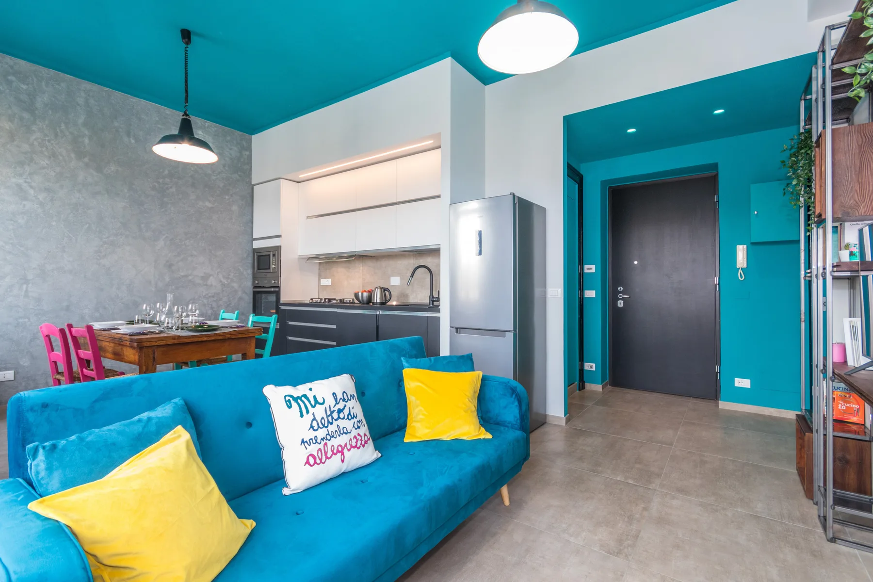 A bright living area with turquoise seating and an open kitchen behind it.