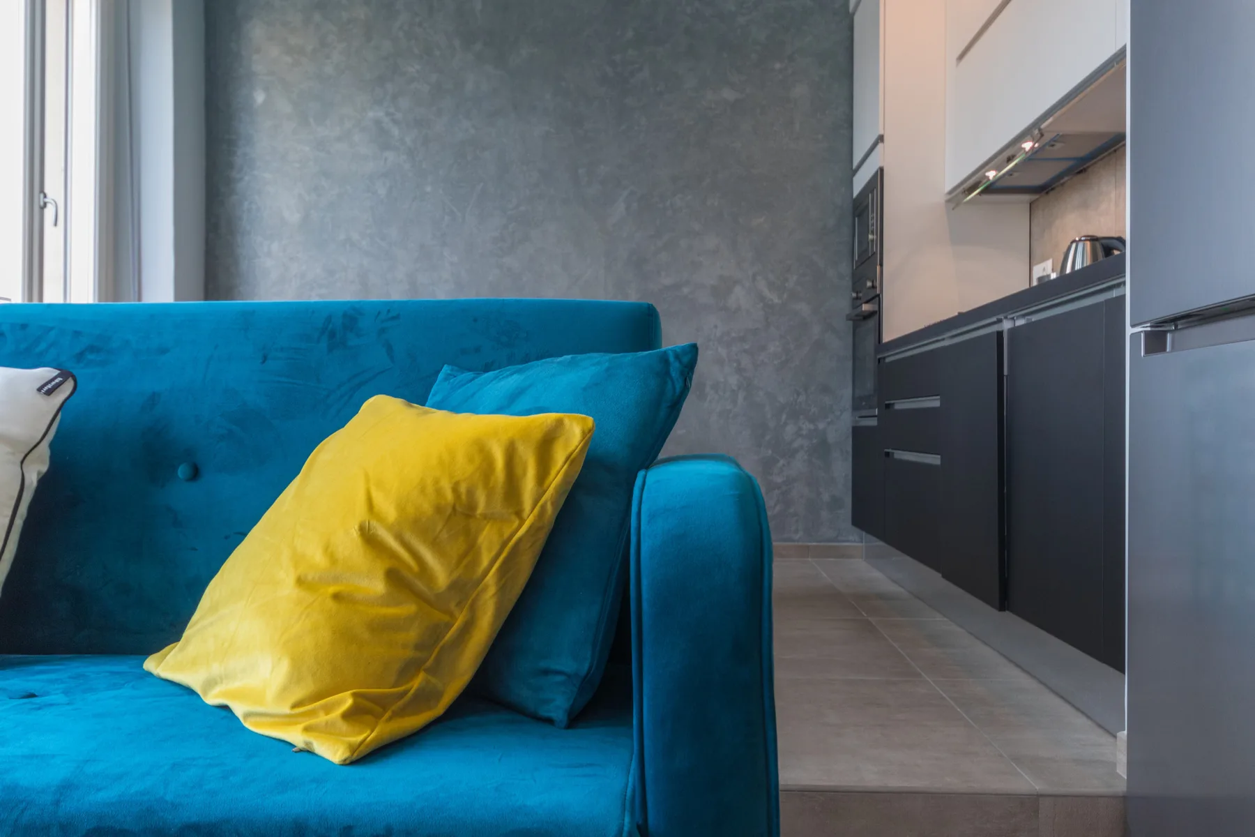 A living room with a blue sofa and a yellow cushion beside the kitchen.