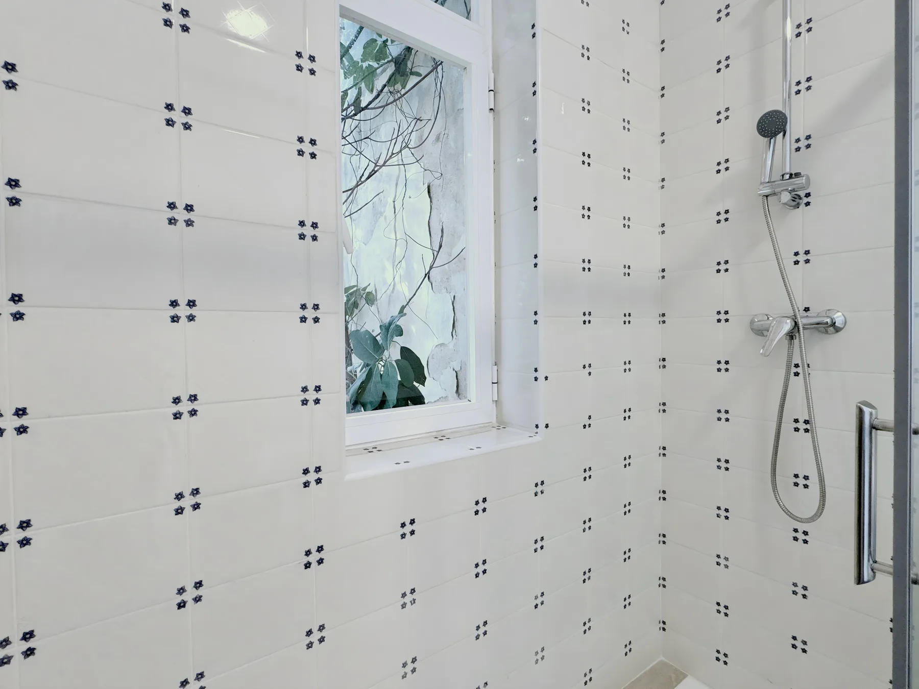 A tiled bathroom shower beside a window with leafy plants outside.