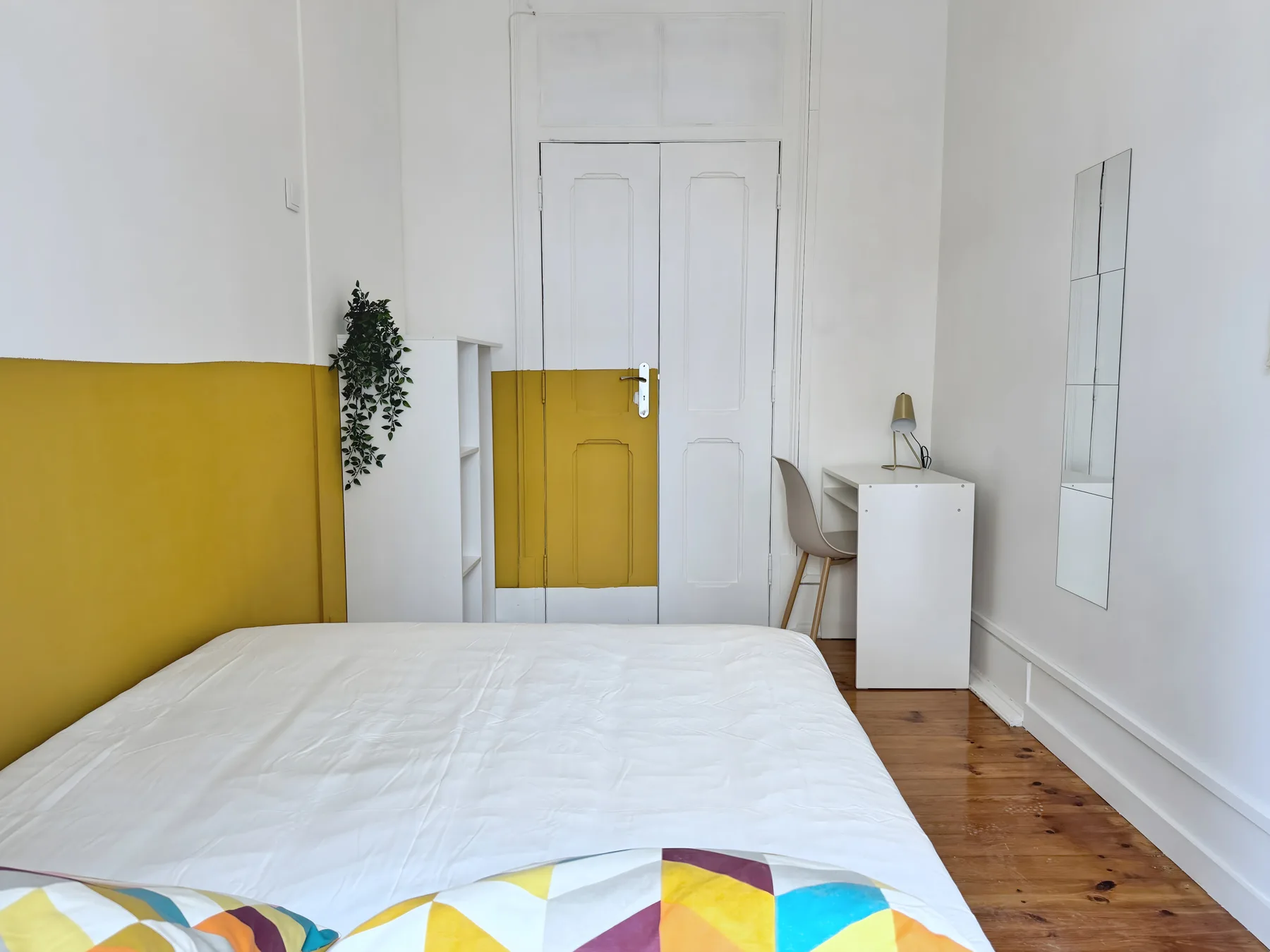 A bedroom with a white bed, mustard-yellow wall panels, and a small desk.