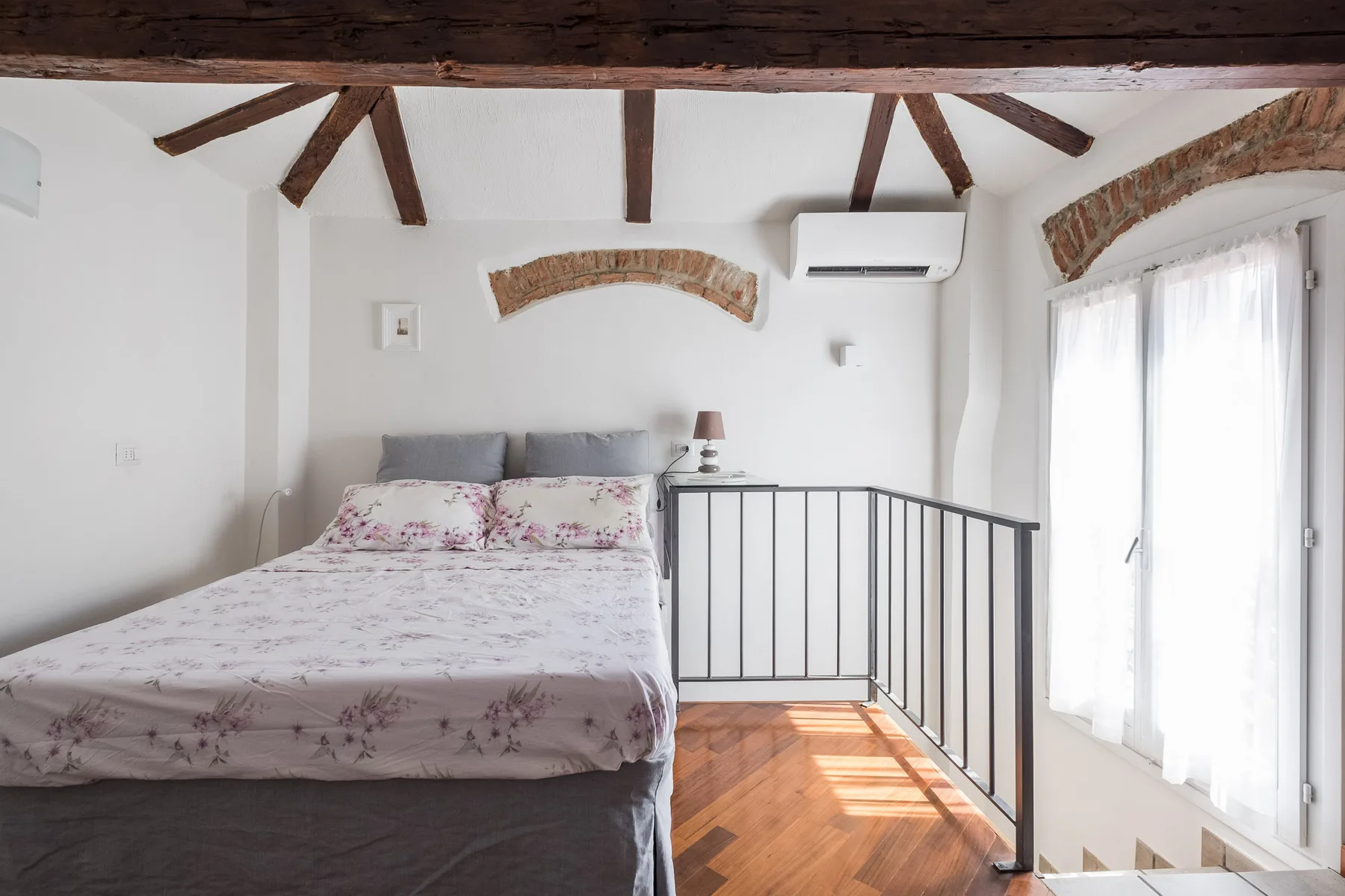 Un dormitorio luminoso con una colcha floral, vigas de techo expuestas y una barandilla junto a la cama.
