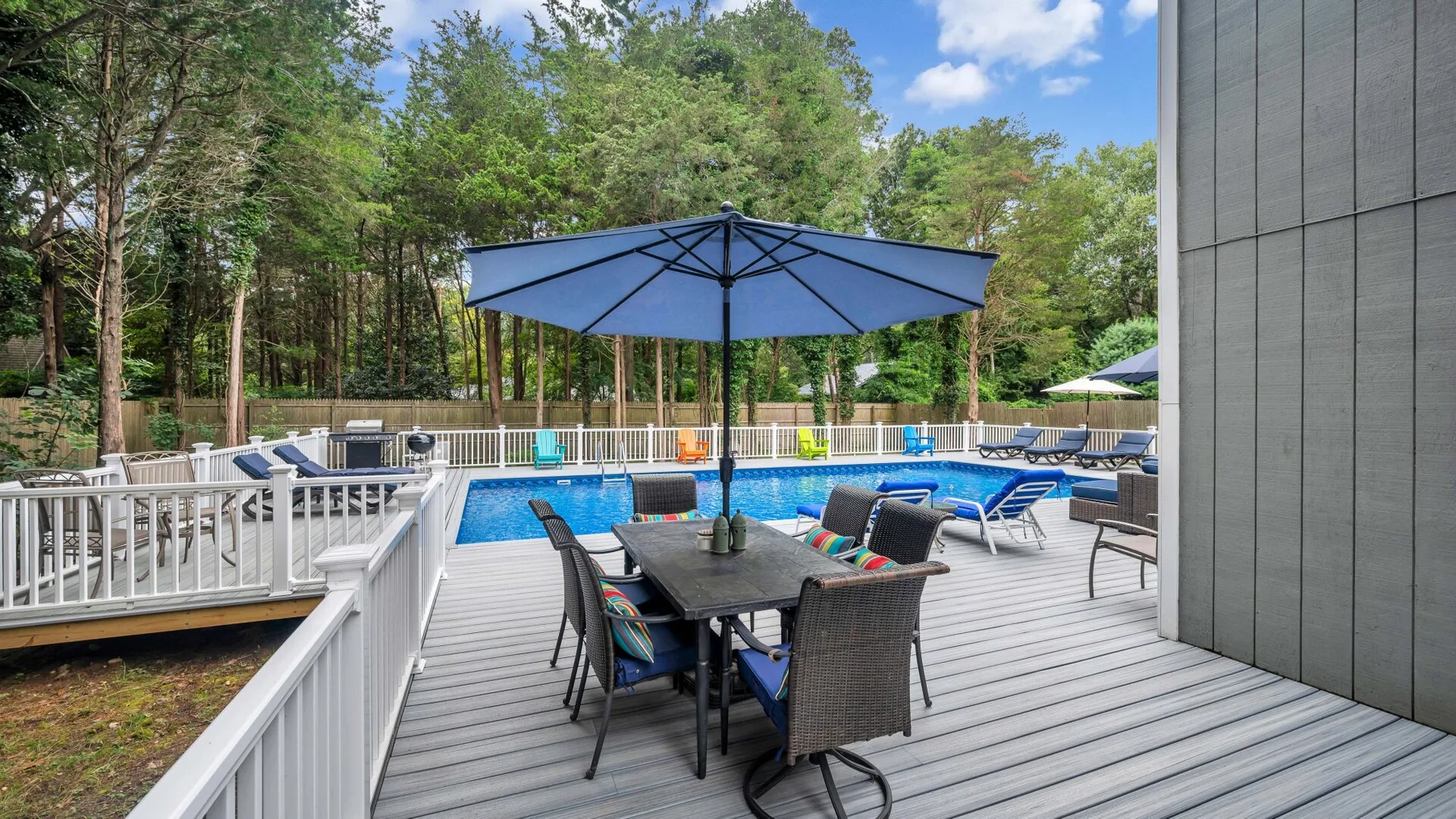 Una terraza junto a la piscina tiene una mesa bajo una sombrilla azul y asientos cerca.
