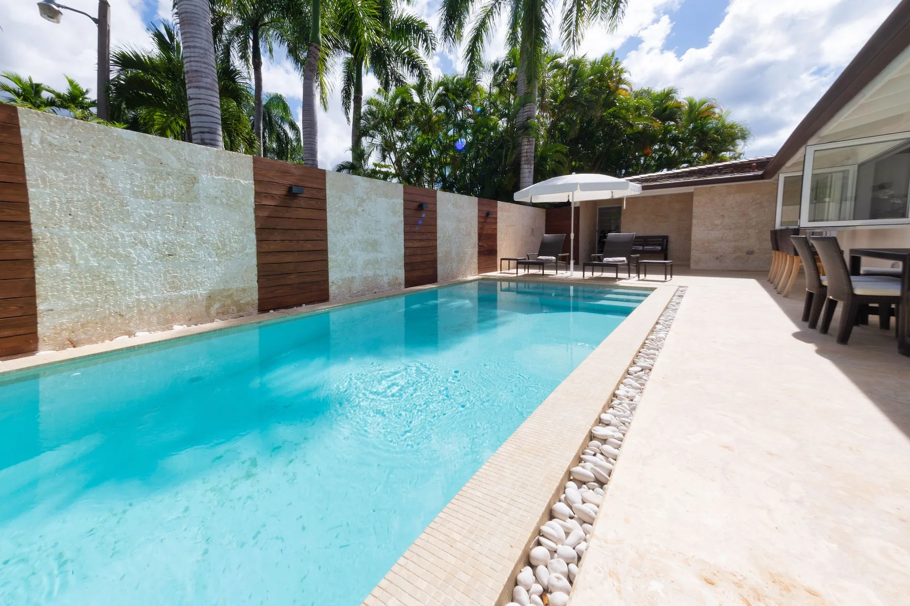 A swimming pool beside a paved patio with outdoor seating and palm trees.