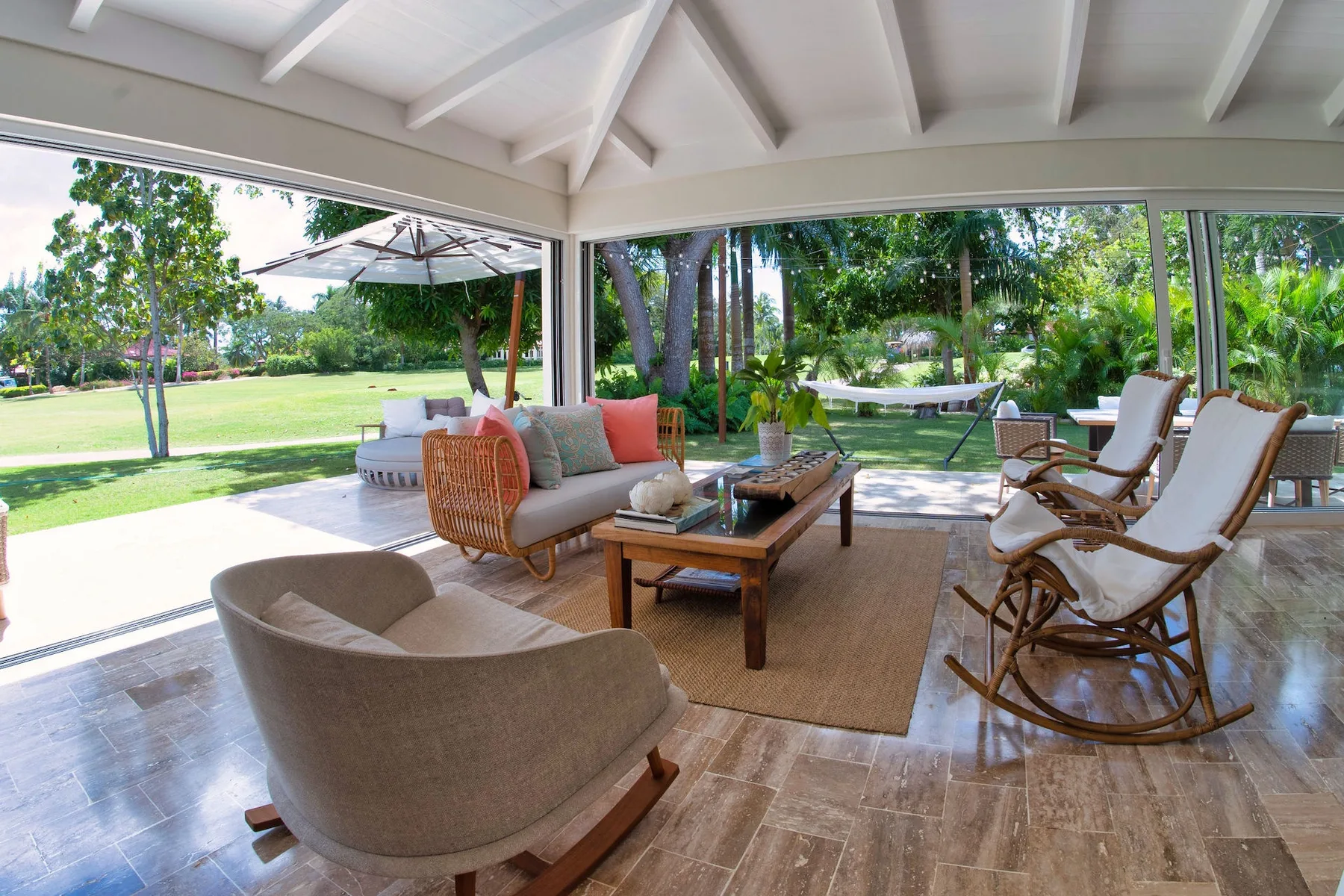 A covered patio lounge opens onto a green garden, with seating and a low table.