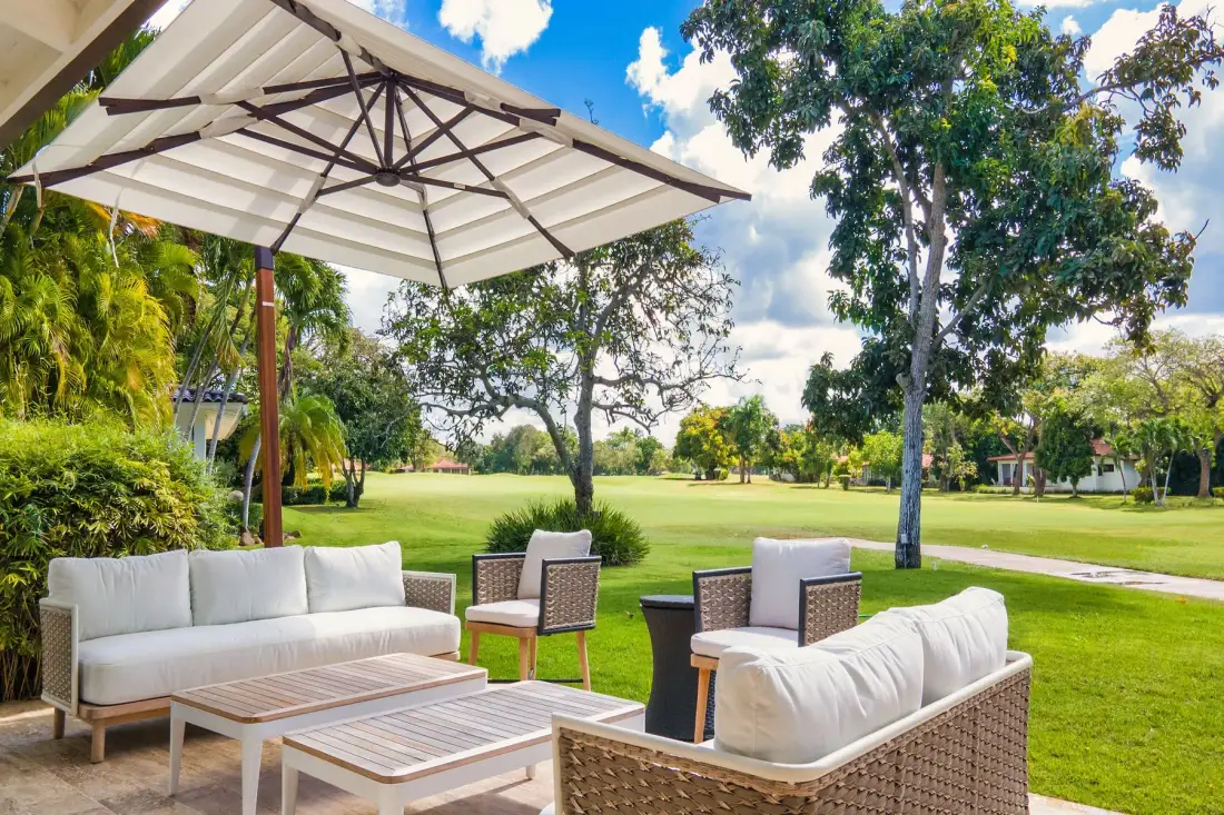 Outdoor seating under a large umbrella overlooks a green lawn and trees.