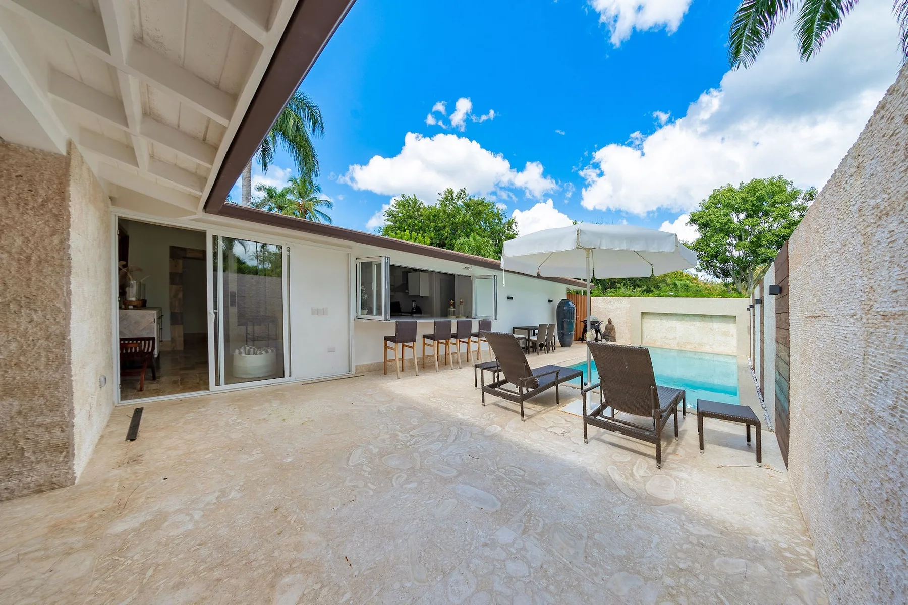 A sunlit outdoor patio with seating and a swimming pool beside the house.