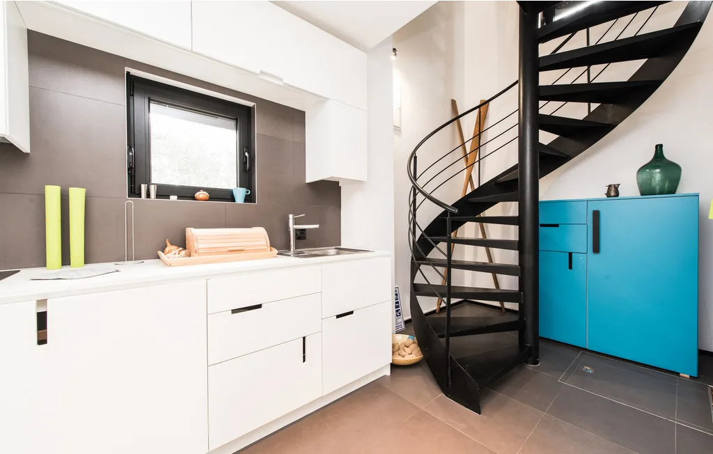 Una pequeña cocina luminosa con armarios blancos está junto a una escalera curva negra y un armario azul.