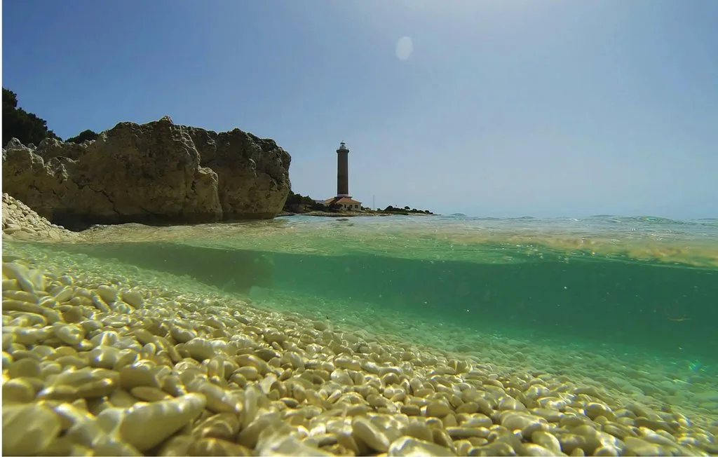 Una costa rocosa y un faro se alzan sobre aguas turquesas y claras.