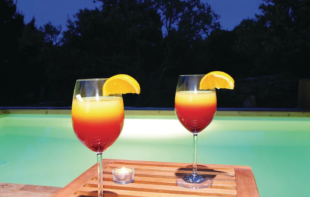 Dos bebidas coloridas están sobre una bandeja de madera junto a una piscina al anochecer.