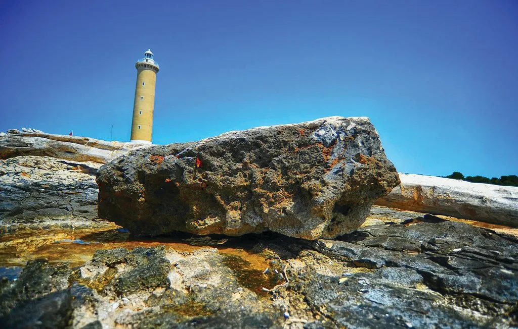 Un faro se alza detrás de una gran roca en una costa rocosa.