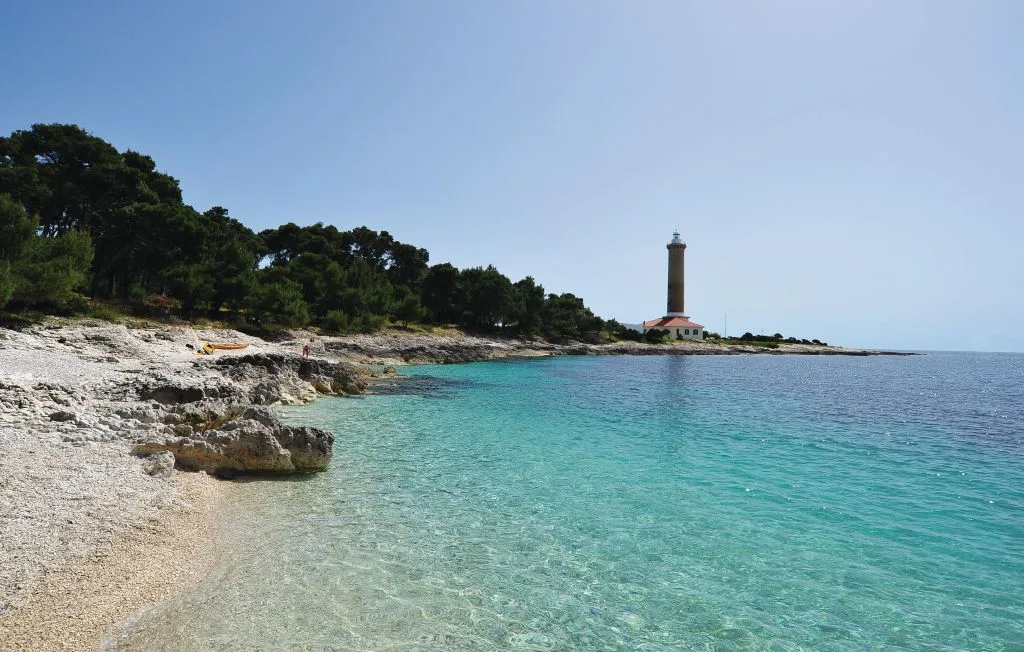 Una playa rocosa junto a aguas turquesas transparentes, con un faro a lo lejos.
