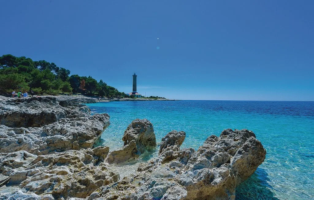 Una costa rocosa bordea aguas azules y claras, con un faro a lo lejos.