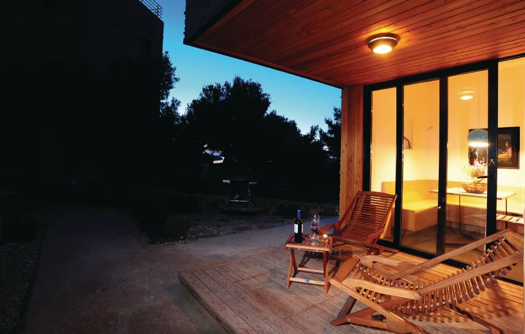 Una terraza de madera con asientos reclinables da a una habitación iluminada al anochecer.