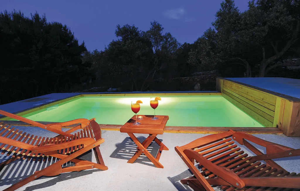 Una piscina exterior iluminada está junto a tumbonas de madera y una mesa pequeña al anochecer.