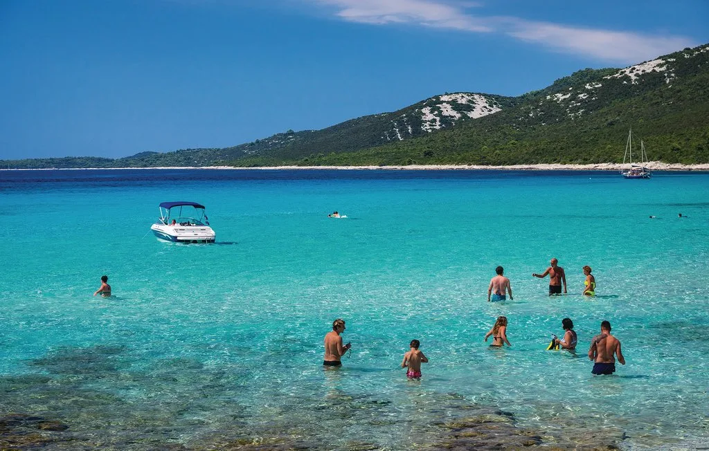 Varias personas caminan por aguas turquesas y claras cerca de un barco, con una costa verde al fondo.