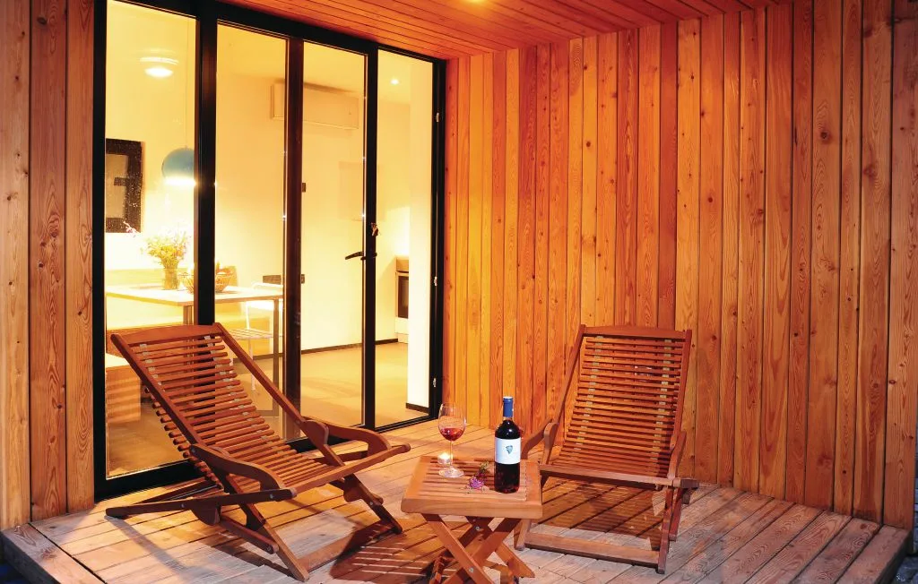 Un patio de madera con dos asientos reclinables y una mesa pequeña junto a puertas de vidrio.