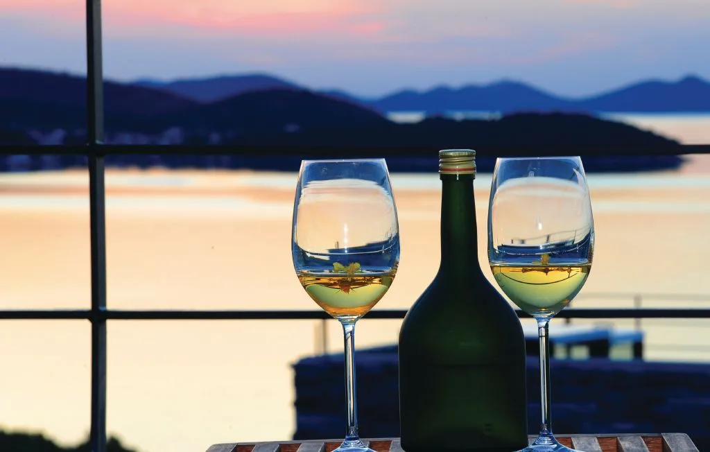 Dos copas y una botella están junto a una ventana con vistas a un lago al atardecer.