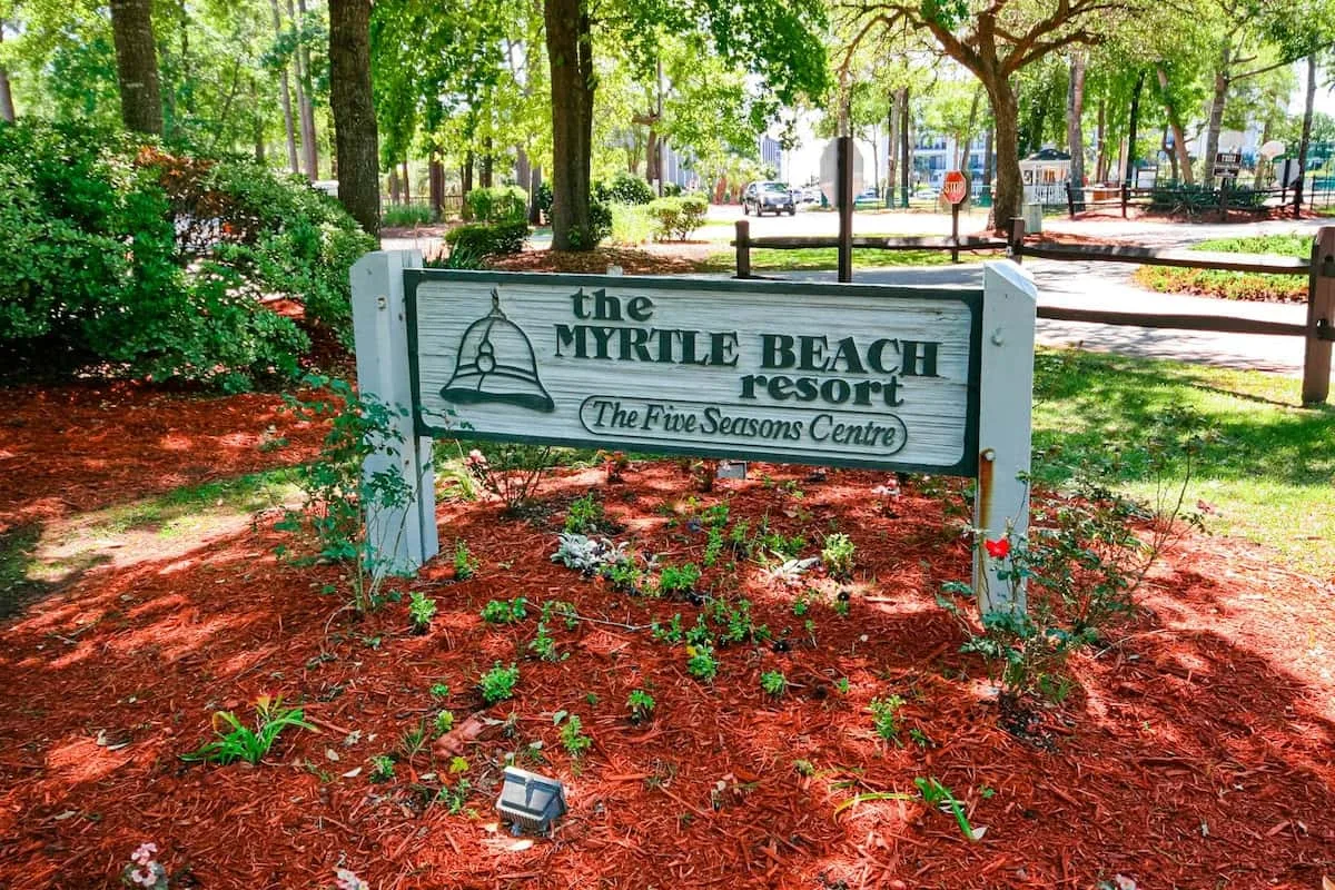 Un letrero de madera de un resort está entre árboles y vegetación ornamental.