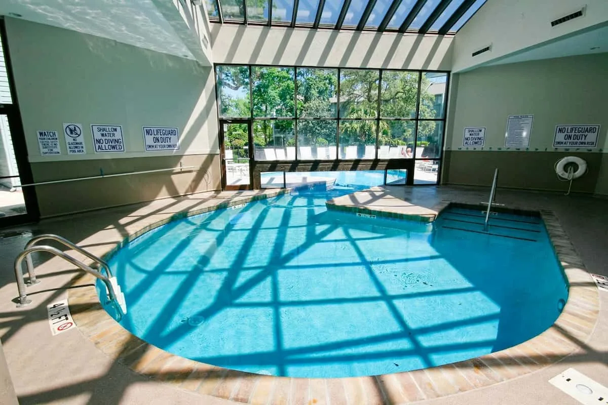 Una piscina cubierta está bajo un techo de cristal, con sombras del sol sobre el agua.