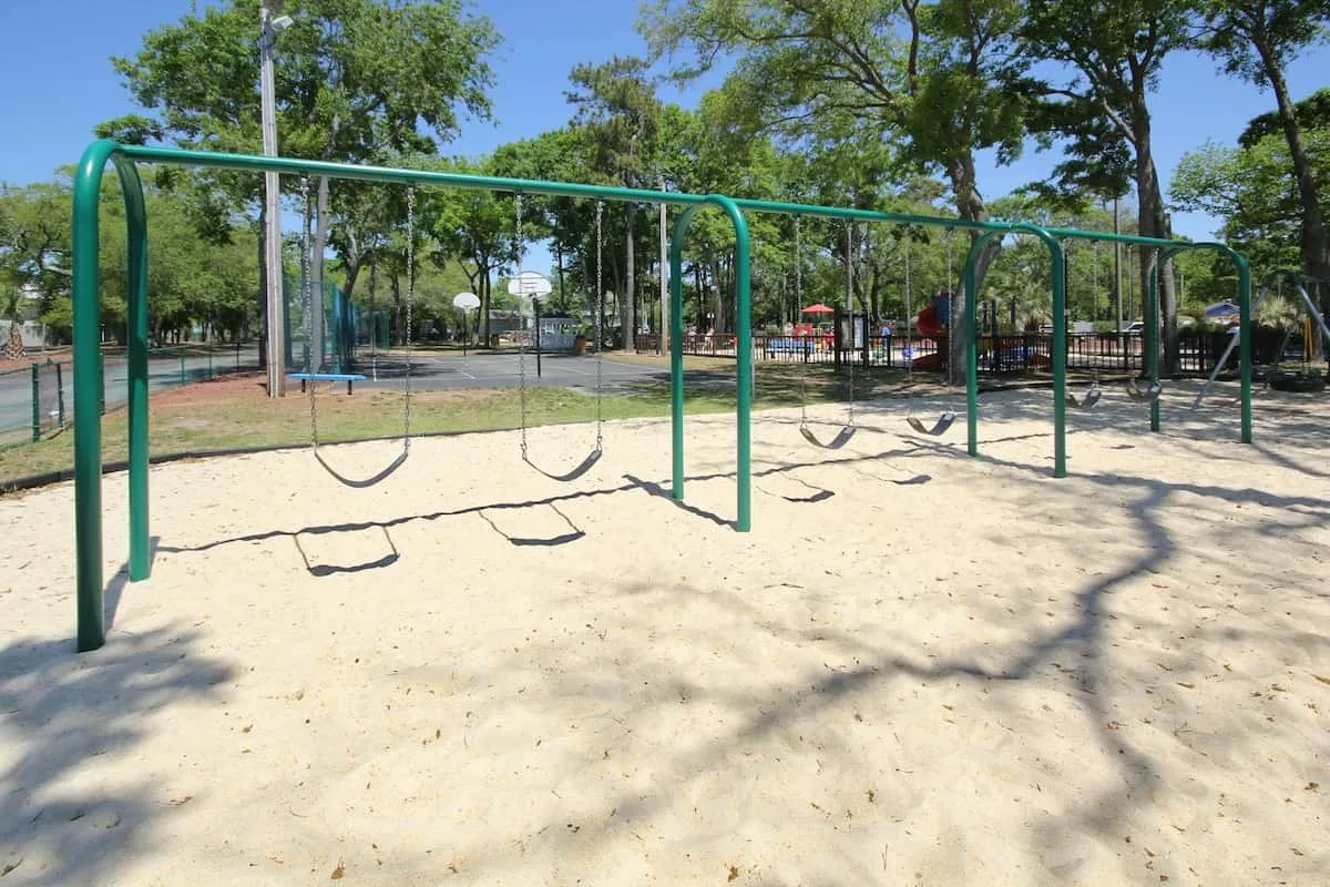 Un columpio de parque infantil se encuentra sobre una zona de arena bajo árboles frondosos.