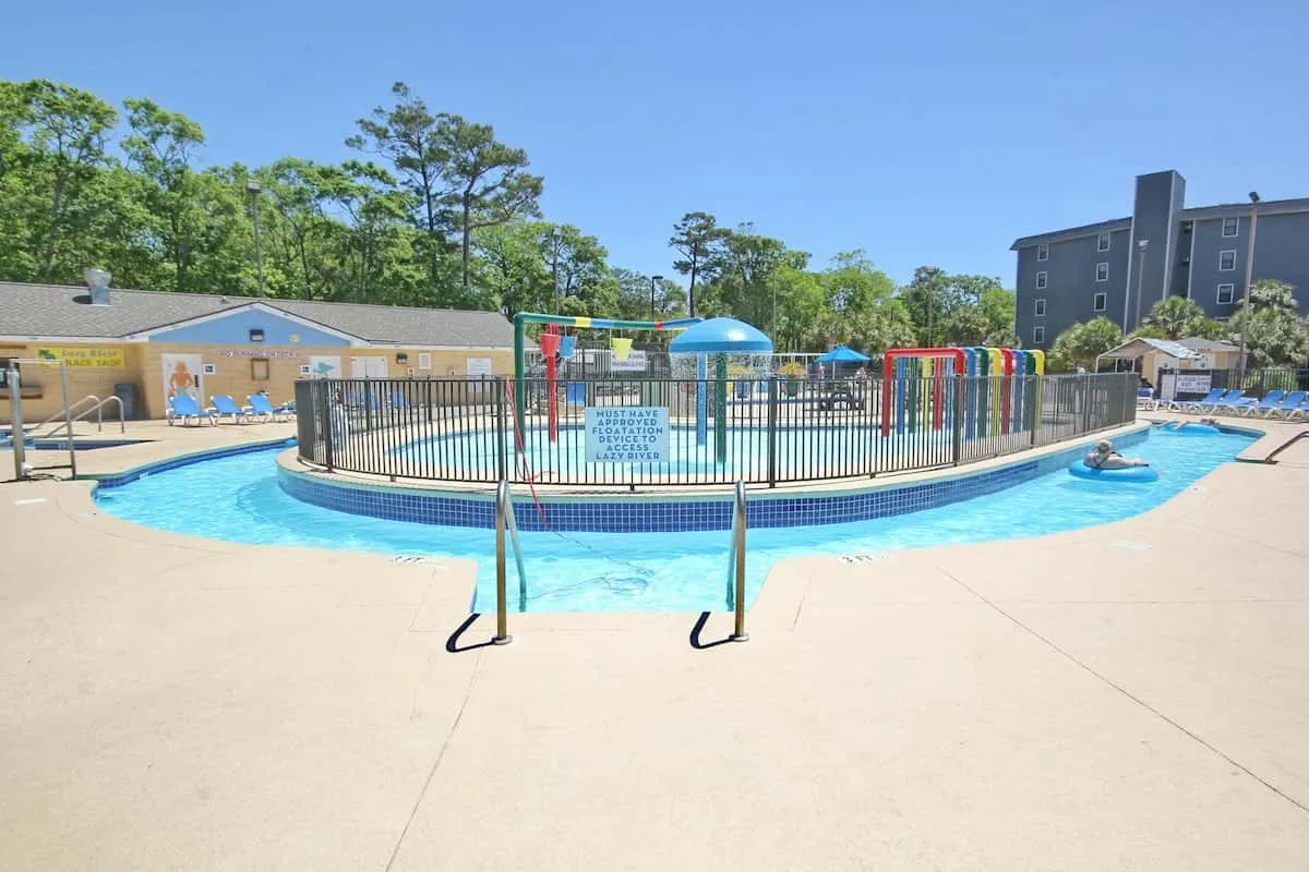 Una piscina al aire libre tiene una zona de juegos cercada y elementos acuáticos coloridos.