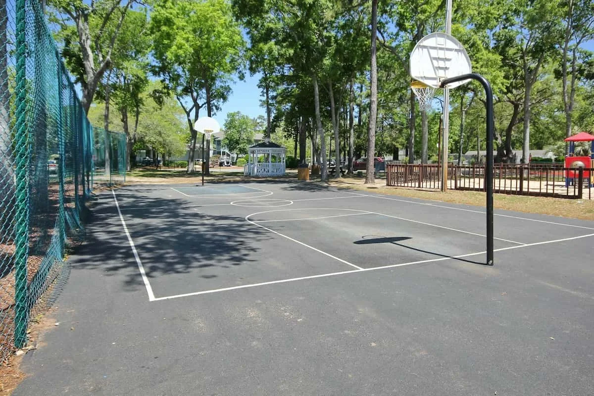 Una cancha de baloncesto al aire libre está entre árboles, con un parque infantil cercado cerca.