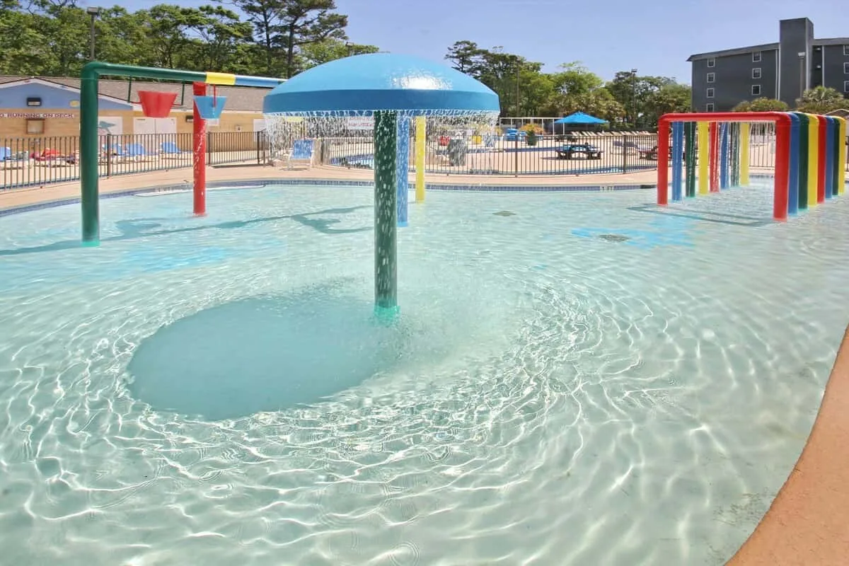 Una piscina infantil al aire libre tiene juegos acuáticos coloridos y una gran fuente con forma de seta.