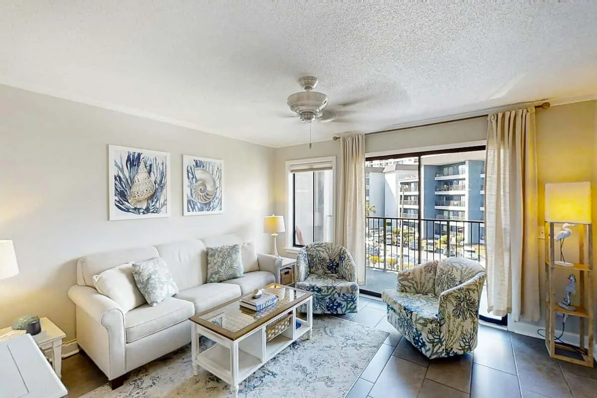 A bright living room with seating, a coffee table, and large windows opening onto a balcony.