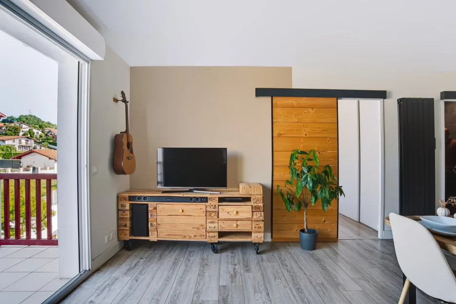 A living and dining area with a television, wooden storage, and sliding doors opening to a balcony.