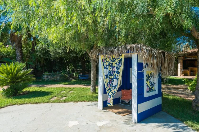 A blue-and-white cabana with patterned curtains sits beneath leafy trees in a garden.