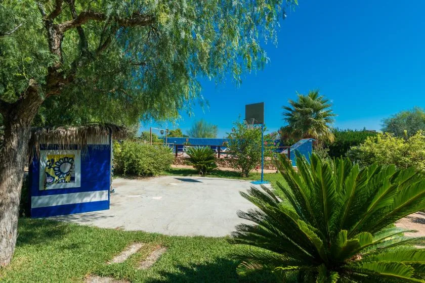An outdoor basketball court sits among trees and shrubs under a clear blue sky.