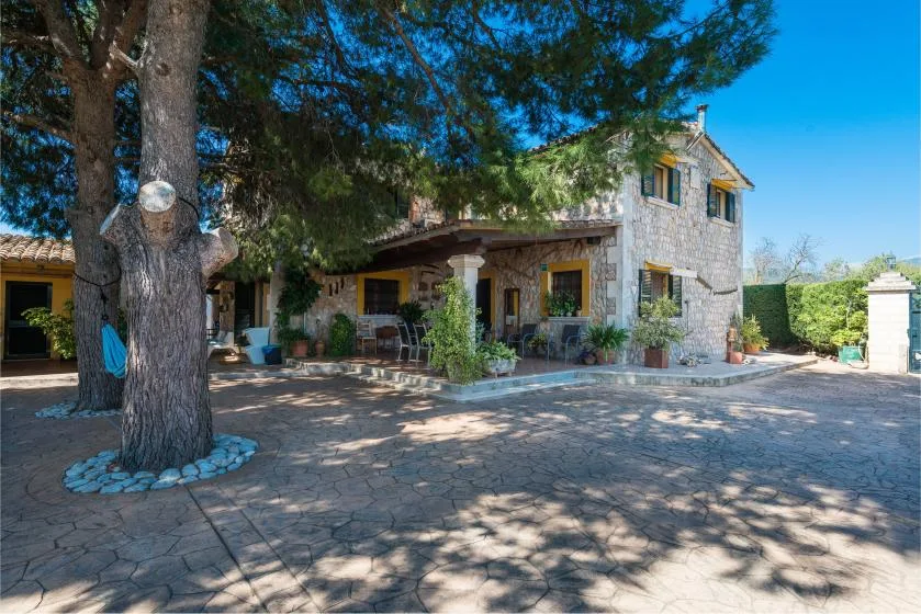 A stone house with a shaded patio and trees in the foreground.