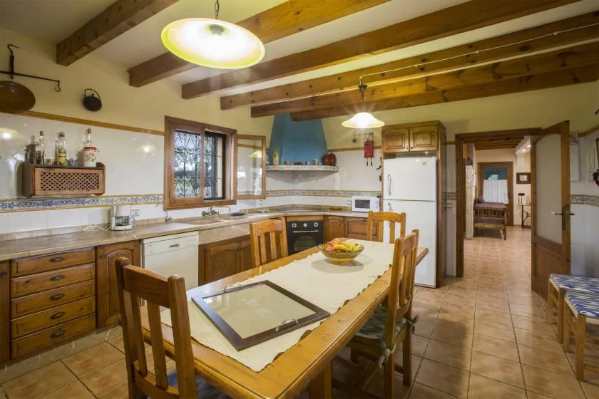 A rustic kitchen with wooden beams, a long dining table, and tiled countertops.