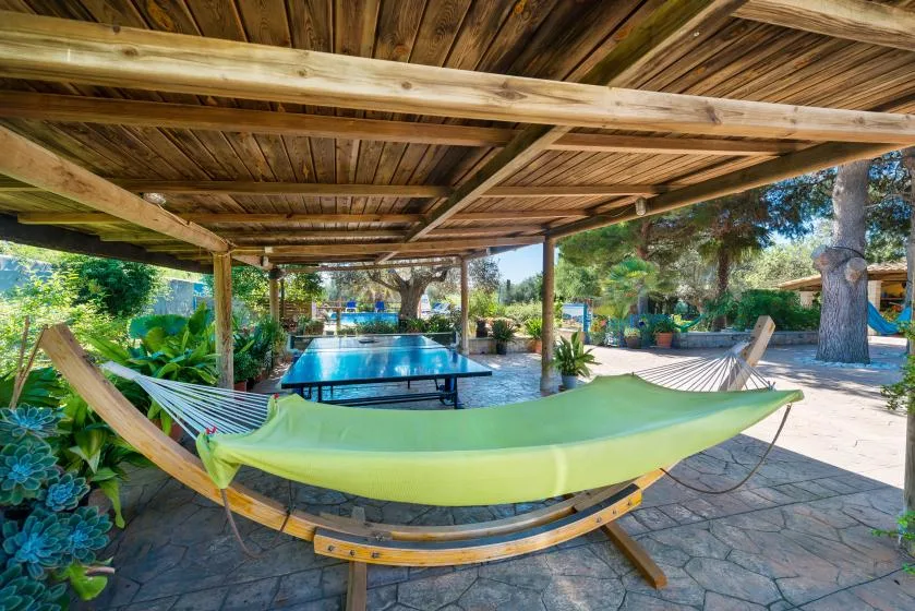 A green hammock hangs under a wooden pergola beside a swimming pool.