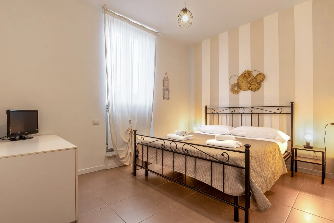 Un dormitorio con una cama de metal, una pared a rayas y una ventana alta con cortinas.
