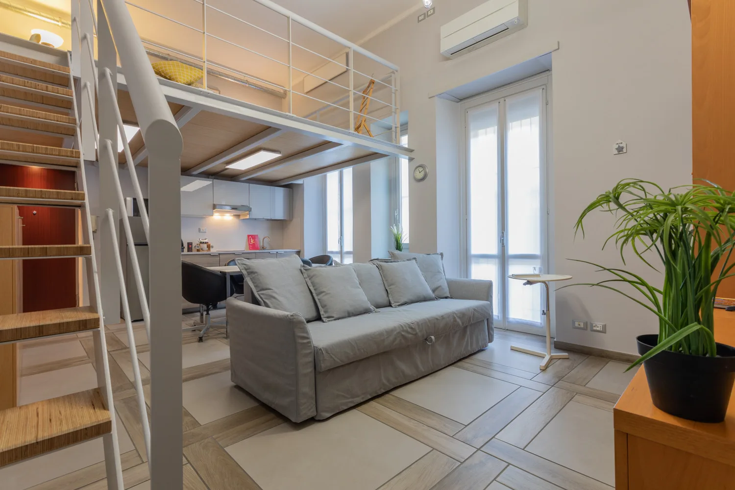 Una luminosa sala de estar con un sofá gris bajo un altillo y una ventana alta.