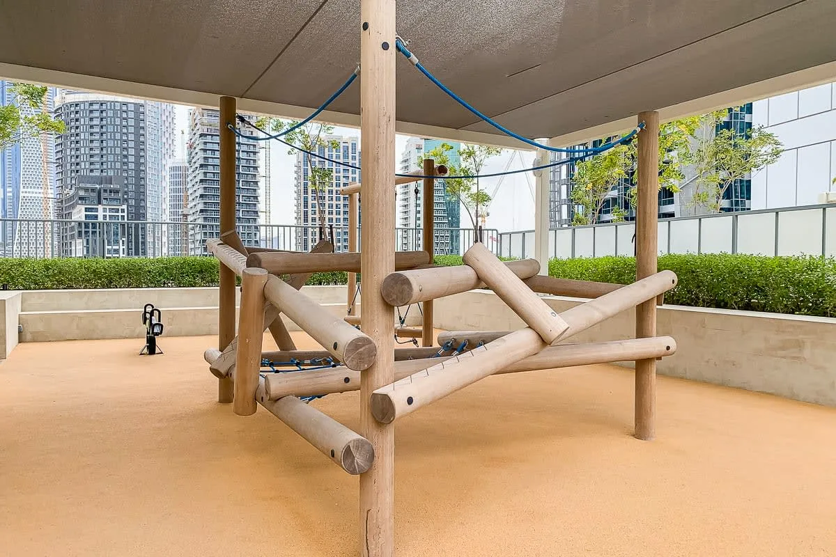 Una zona de juegos exterior cubierta tiene una estructura de madera para trepar y un espacio pavimentado abierto.