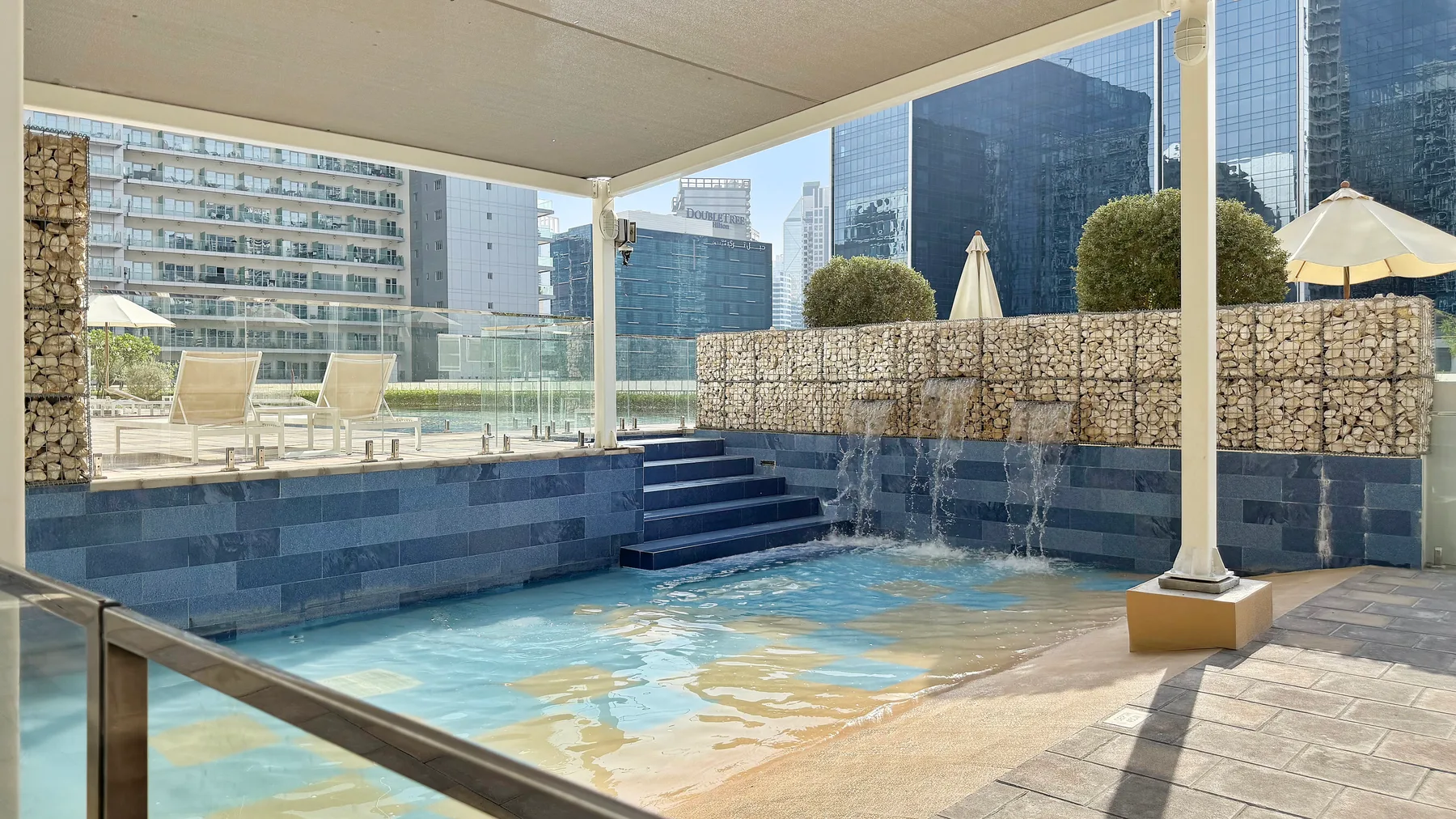 Una pequeña piscina exterior está junto a un muro de piedra, con edificios urbanos al fondo.