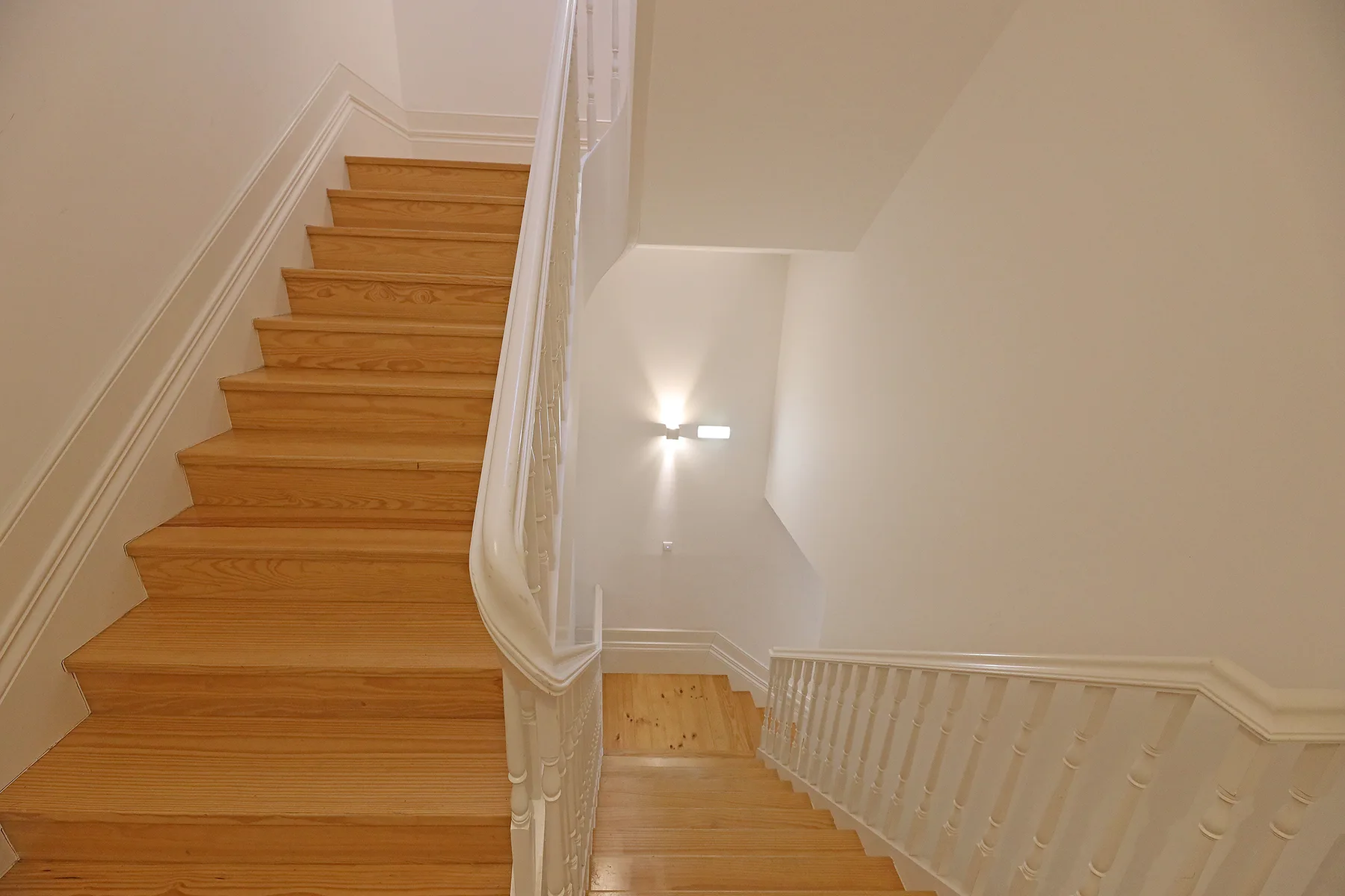 Una escalera de madera desciende por un interior luminoso con barandillas blancas.