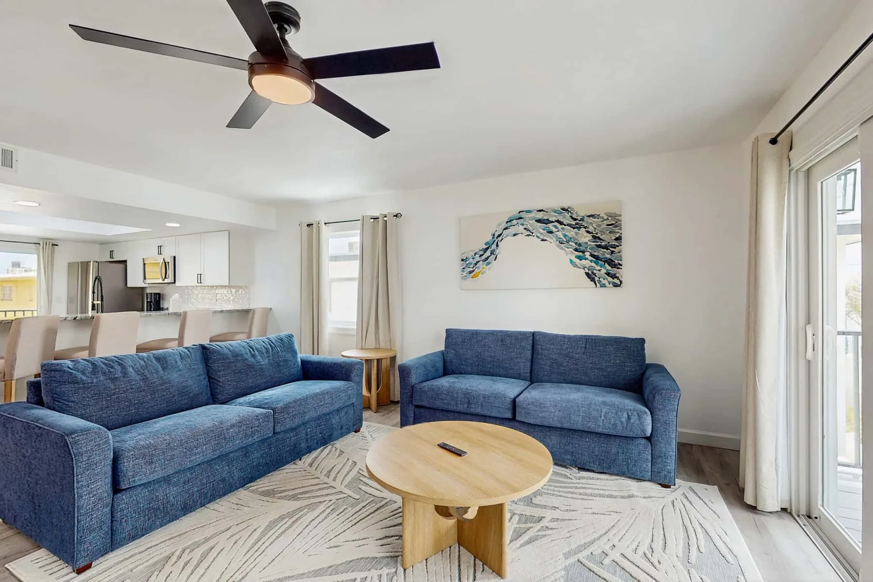 A bright living room with blue seating, a round coffee table, and an open kitchen.