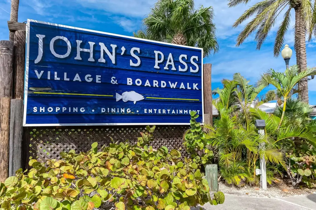 A blue John’s Pass Village and Boardwalk sign stands among tropical plants.