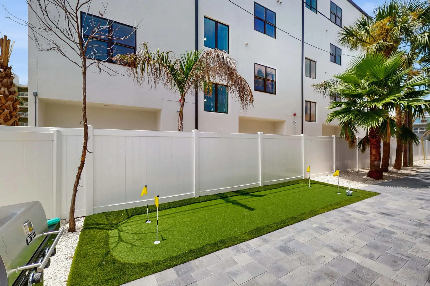 An outdoor putting green sits beside a white fence and paved walkway.