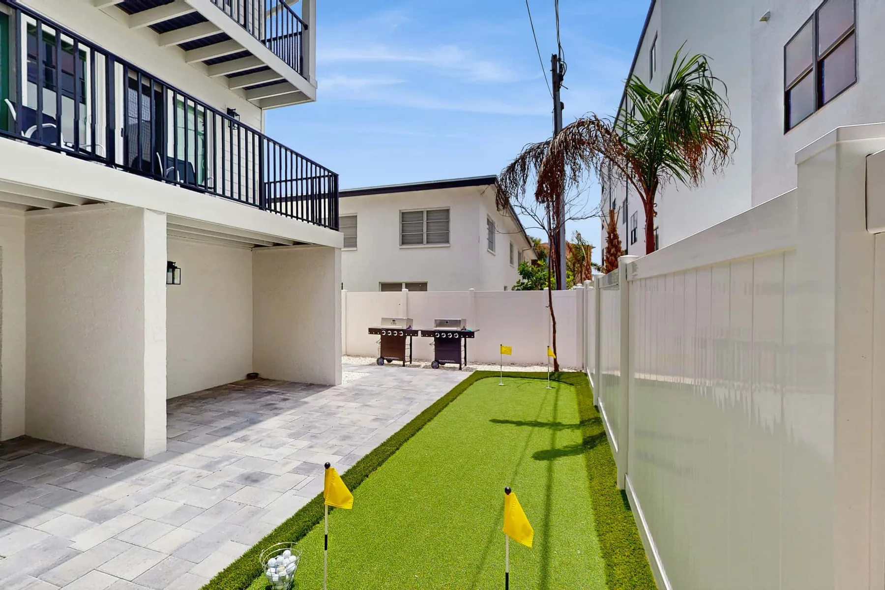 An outdoor patio beside a narrow strip of green turf, with buildings and a fence.