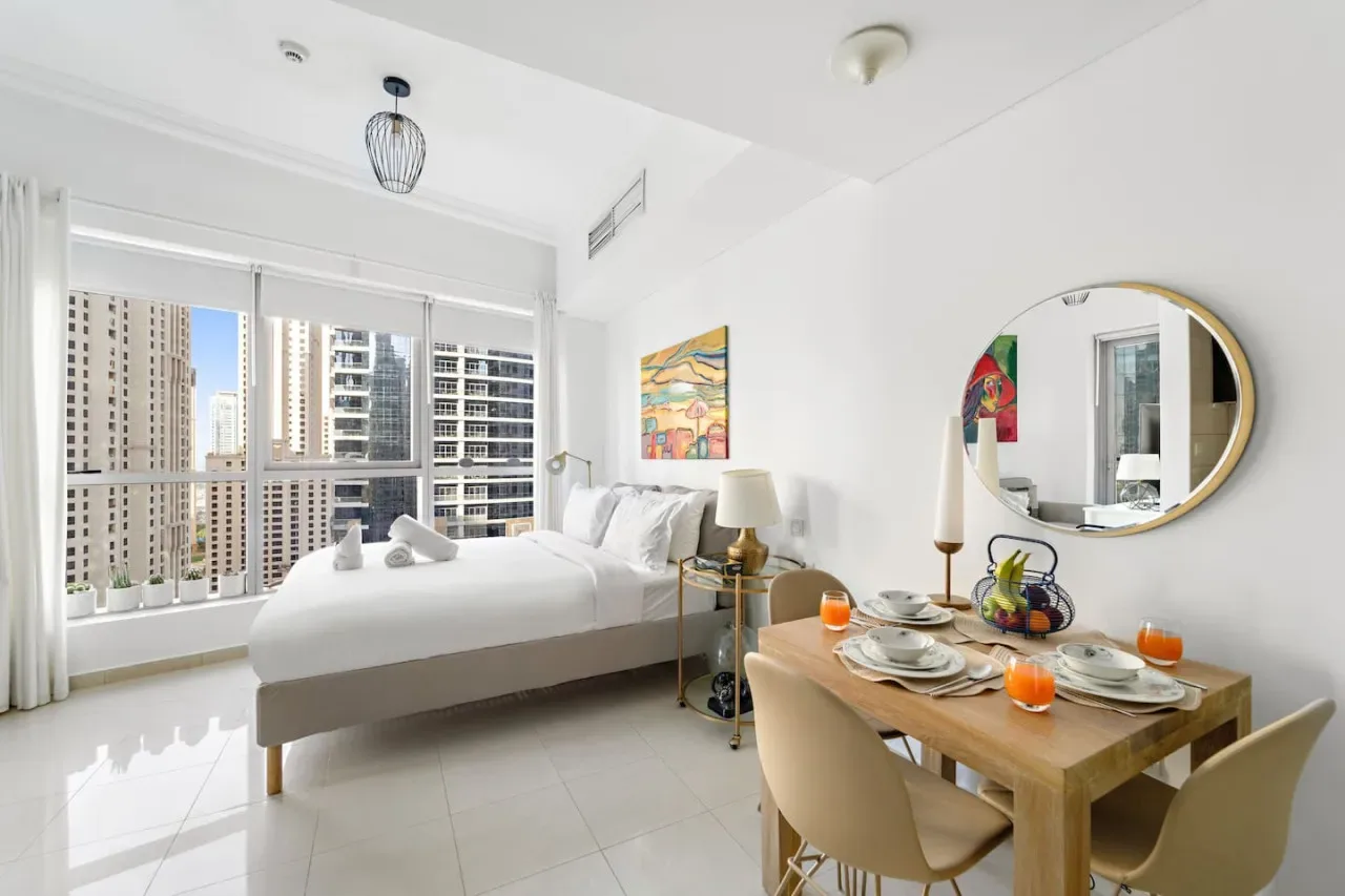 Un dormitorio luminoso con una cama junto a una mesa de comedor y grandes ventanales.