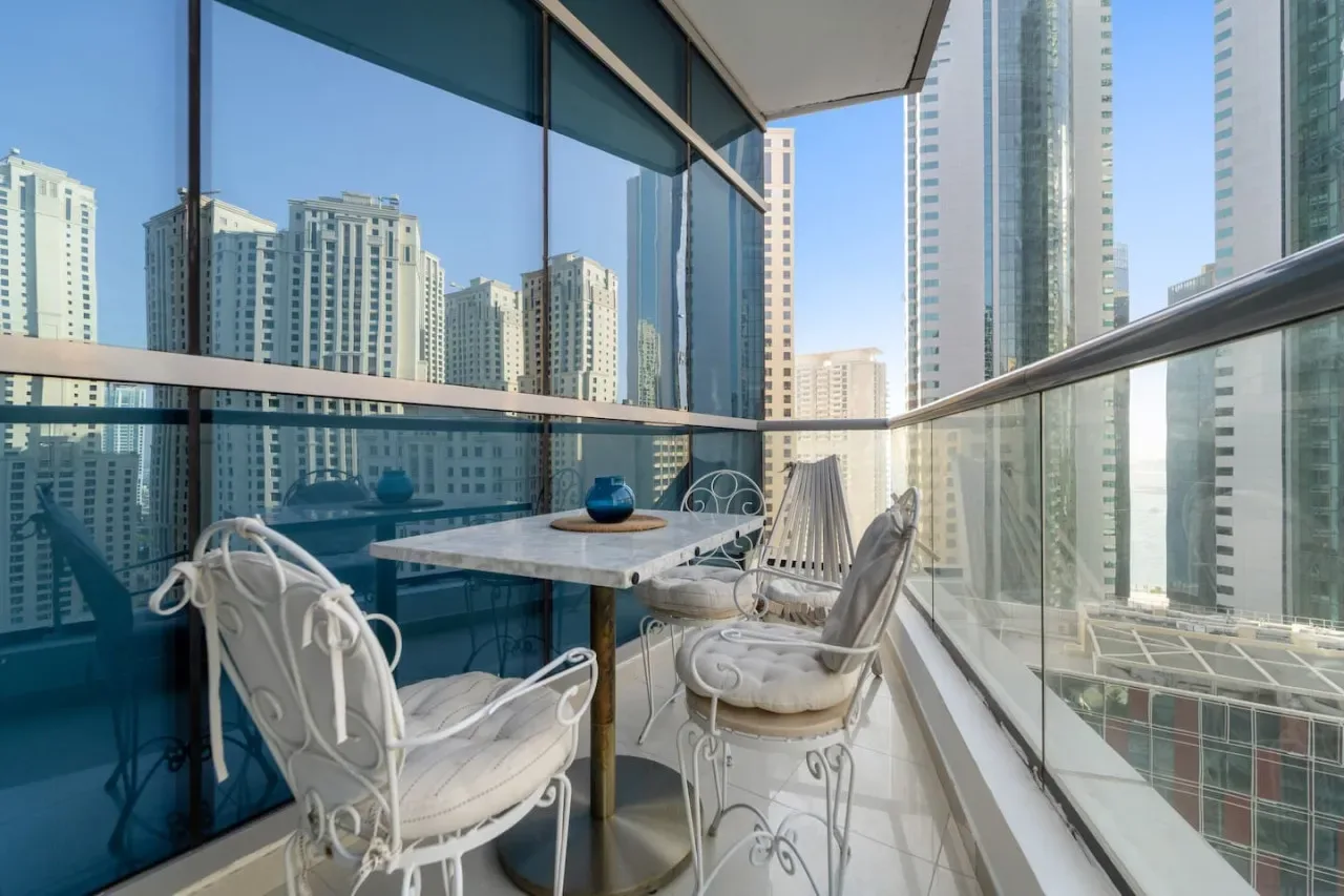 Un balcón acristalado con asientos y una mesa pequeña tiene vistas a edificios altos cercanos.