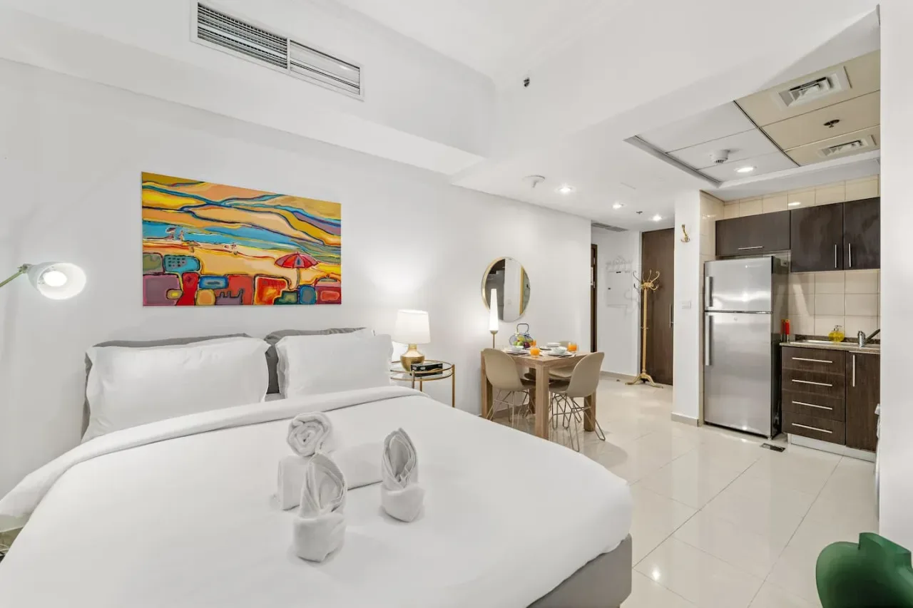 Un dormitorio luminoso con ropa de cama blanca se abre a una cocina y comedor compactos.